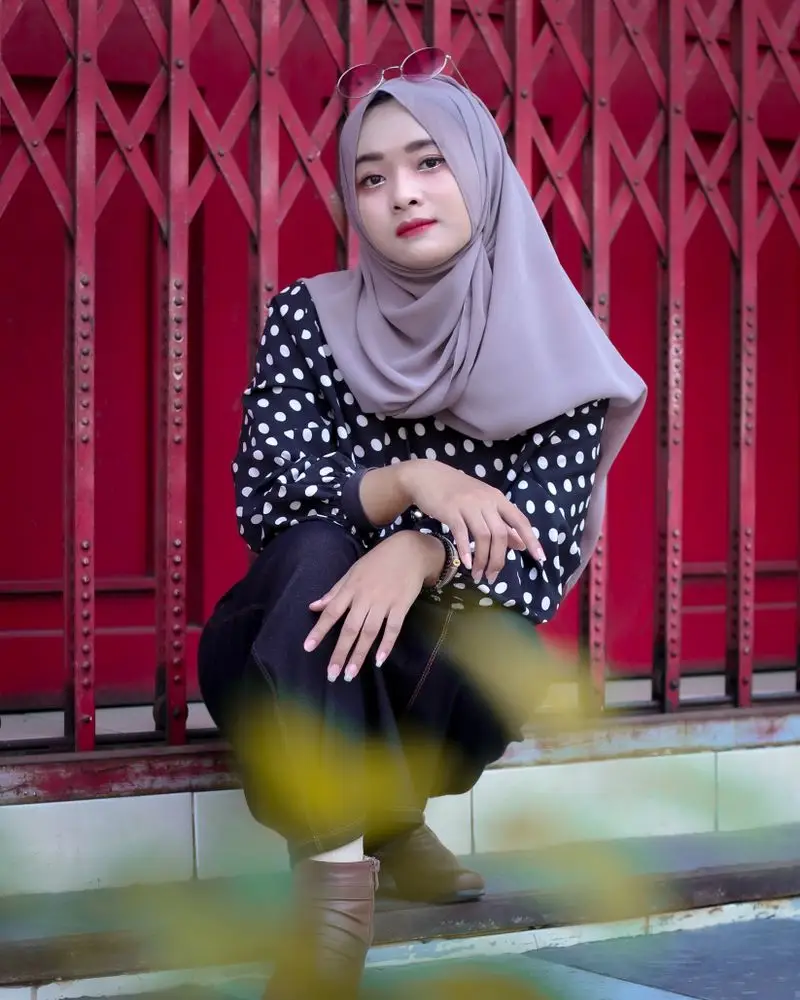 Blus polkadot dan hijab abu di depan pintu besi merah